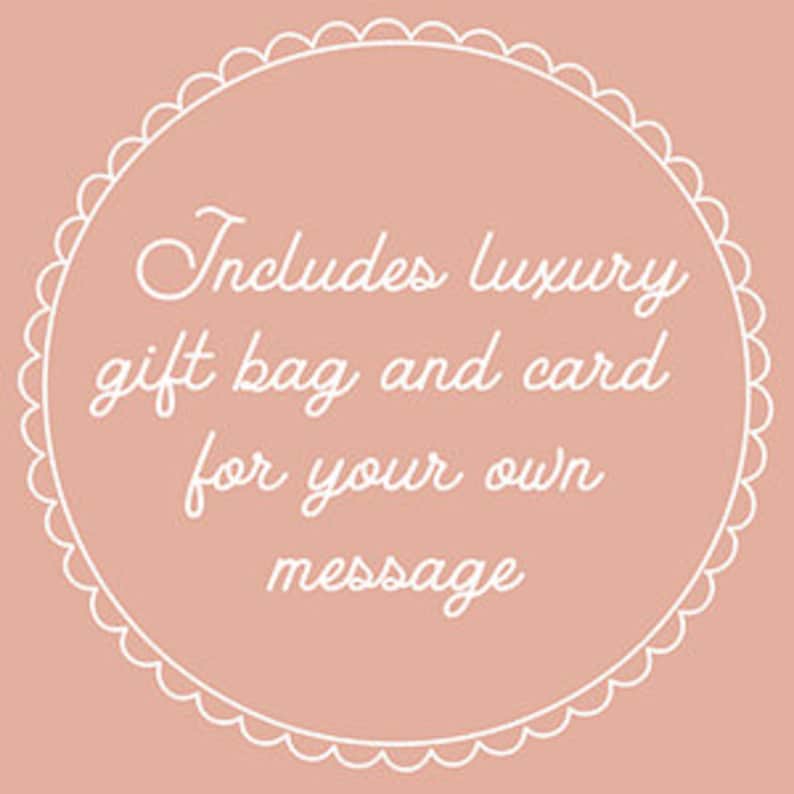 Puede incluir: Un c&iacute;rculo blanco con un borde festoneado sobre un fondo rosa claro. El texto "Includes luxury gift bag and card for your own message" est&aacute; escrito en blanco en cursiva.