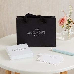 Puede incluir: Una bolsa de regalo negra con un logotipo blanco que dice "Angel & Dove". La bolsa est&aacute; sobre una mesa blanca con una caja blanca que tambi&eacute;n dice "Angel & Dove" y una tarjeta blanca con el mismo logotipo.