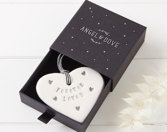 May include: A white ceramic heart ornament with the text "FOREVER LOVED" and small heart cutouts, suspended by a black and white gingham ribbon. It is presented inside a black sliding gift box featuring the text "ANGEL & DOVE" and star icons.