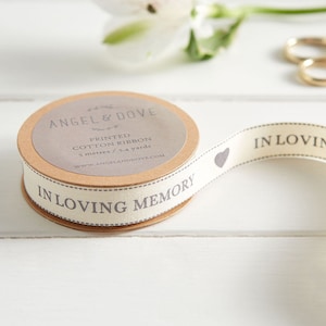 May include: A roll of beige cotton ribbon with the text "In Loving Memory" printed in grey. The ribbon is 5 metres or 5.4 yards long and is packaged on a brown cardboard spool with the brand name "Angel & Dove" printed on it.