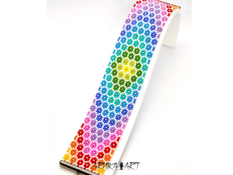 以下が含まれることがあります： 小さな多色のビーズを六角形のフラワーパターンで並べた幅広のブレスレット。赤、オレンジ、黄色、緑、青、紫と虹色のスペクトルで色が変化しています。ARNIKA ARTという文字がプリントされています。