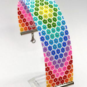 以下が含まれることがあります： 赤、オレンジ、黄、緑、青、紫のビーズが白地の上で鮮やかな虹色のグラデーションを描く、花柄の繰り返し模様が特徴の幅広のビーズブレスレット。ブレスレットには銀色の金属製エンドバーとロブスタークラスプが付いています。