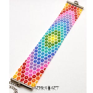 以下が含まれることがあります： 小さな花形のビーズで構成された鮮やかな虹色のグラデーション模様の幅広ビーズブレスレット。赤、オレンジから黄色、緑、青、紫へと色が変化するデザインです。銀色の金属製エンドバーとチェーンクラスプが付いています。