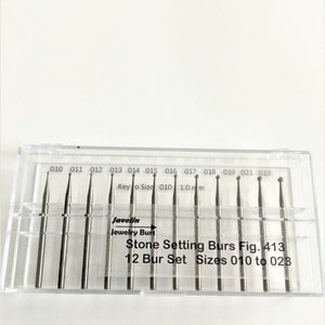 May include: A clear plastic case containing a set of 12 stone setting burs. The burs are silver-colored and vary in tip shape and size, ranging from 010 to 023. The case is labeled "Javelin Jewelry Burs".