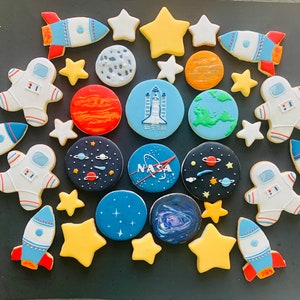 May include: A collection of decorated sugar cookies with space-themed designs, including rockets, astronauts, planets, and stars. The cookies are decorated with colorful icing and details.