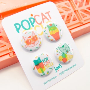 May include: Set of four 1-inch button badges featuring a cat wearing sunglasses, a cat eating ice cream, a cat eating a popsicle, and the text "Pop Cat Sicle". The badges are white with a colourful design and are displayed on a white card with the text "Pop Cat Sicle" and "Jaws Boutique".