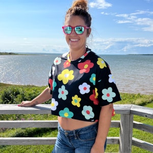 May include: A woman wearing a black cropped t-shirt with a colourful floral print. The shirt has short sleeves and a round neckline. The woman is wearing blue jeans and has her hair pulled back in a bun. She is smiling and wearing sunglasses.