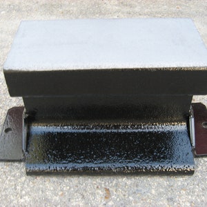 May include: A black metal anvil with a gray top surface. The anvil has a rectangular base with two metal brackets on the sides.