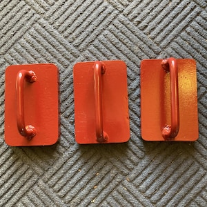 May include: Three red rectangular blocks with curved metal handles. The blocks are evenly spaced and appear to be made of painted metal. The background is a textured, dark blue-gray surface.