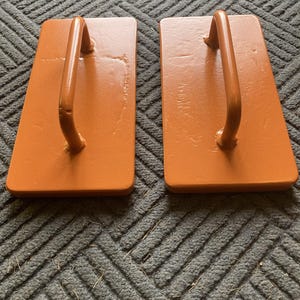 May include: Two orange-brown wooden blocks with curved metal handles. The blocks are rectangular and have a slightly worn appearance. The handles are attached to the top of each block, creating a sturdy grip. The blocks are placed on a gray textured surface.