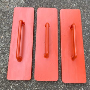 Puede incluir: Tres llana de mano rectangulares rojas con asas naranjas. Estas herramientas se utilizan para alisar y acabar superficies de hormigón o yeso. Las asas están curvadas para facilitar el agarre.