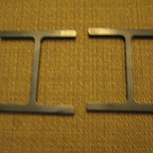 May include: Two metal T-shaped pieces, each with a flat top and a vertical stem. The pieces are identical and are positioned side-by-side.