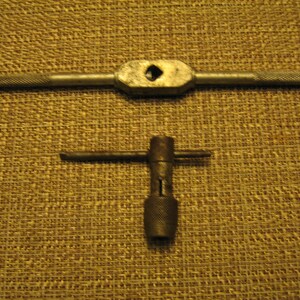 May include: Two metal tap wrenches, one with a long handle and a diamond-shaped opening, and another with a T-shaped handle. Both tools have textured grips for a secure hold. The tools are likely used for threading or rethreading holes.