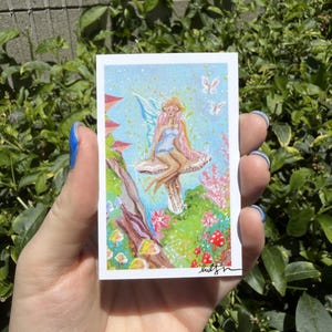 May include: A rectangular art print featuring a painting of a fairy with pink hair and blue wings sitting on a large mushroom. The scene includes a blue sky, small white butterflies, red mushrooms, and pink flowers. The artist's signature is in the corner.