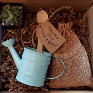 May include: A small blue watering can, a burlap bag, a wooden spoon, a succulent plant in a pot, and a brown paper tag with the text "Thank You" in a gift box filled with brown shredded paper.