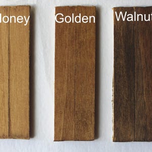 Może przedstawiać: Pięć prostokątnych próbek drewna w różnych kolorach bejcy: Naturalny, Miodowy, Złoty, Orzechowy i Wiśniowy. Każda próbka ma u góry nadrukowaną na biało nazwę koloru. Słoje drewna są widoczne w każdej próbce.