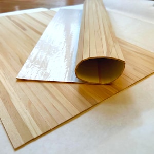 Puede incluir: Láminas de chapa de madera de color claro, una plana y la otra enrollada. La chapa está hecha de finas tiras de madera, creando un patrón a rayas. Una lámina de material transparente y brillante cubre parcialmente la madera.