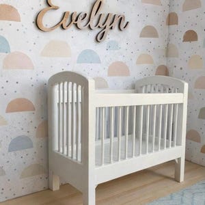 May include: A white crib with vertical slats and a matching wooden name sign that reads "Evelyn" is displayed in a nursery. The walls are decorated with a pastel-colored, semi-circle pattern. A light blue rug is on the floor.