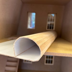 Może przedstawiać: Miniaturowe wnętrze domku dla lalek z tunelopodobną konstrukcją z jasnego drewna. Tunel jest cylindryczny z białym wnętrzem. Domek dla lalek ma okna i schody, wszystko w neutralnej palecie kolorów.