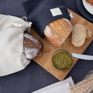 Peut inclure: Deux sacs à pain en lin, un blanc et un bleu foncé, avec un pain dans chacun. Le sac blanc porte une étiquette qui dit "TILL". Le sac bleu a une étiquette carrée blanche. Il y a une planche à découper en bois avec un pain, deux tranches de pain et un petit bol en verre de pesto vert.