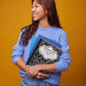 Könnte beinhalten: Eine Person in einem hellblauen Pullover und Jeans hält einen Stapel Schreibhefte. Das oberste Notizbuch hat einen schwarz-weiß gesprenkelten Einband mit dem Aufdruck "COMPOSITION BOOK".