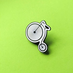 A penny farthing enamel pin against a green background. The pin shows the old-style bicycle in black surrounded by a white recessed border.