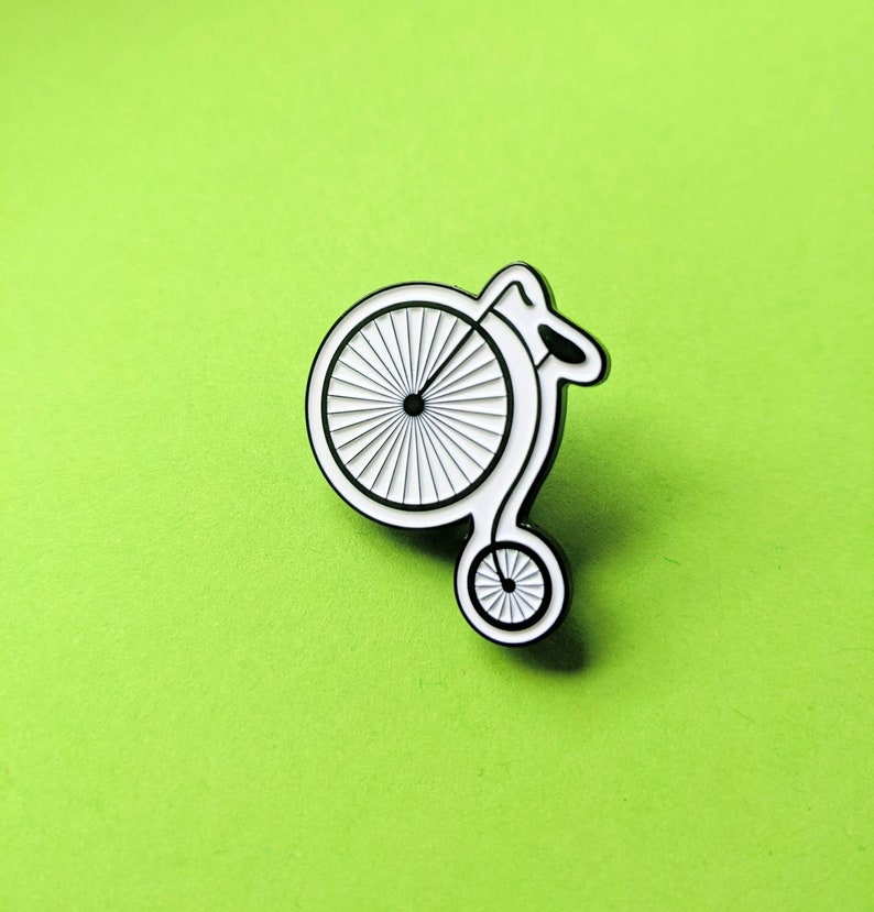 A penny farthing enamel pin against a green background. The pin shows the old-style bicycle in black surrounded by a white recessed border.