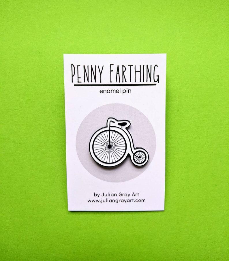 The penny farthing pin pinned onto a backing card, which is white and black and says: Penny Farthing Enamel Pin by Julian Gray Art www.juliangrayart.com.