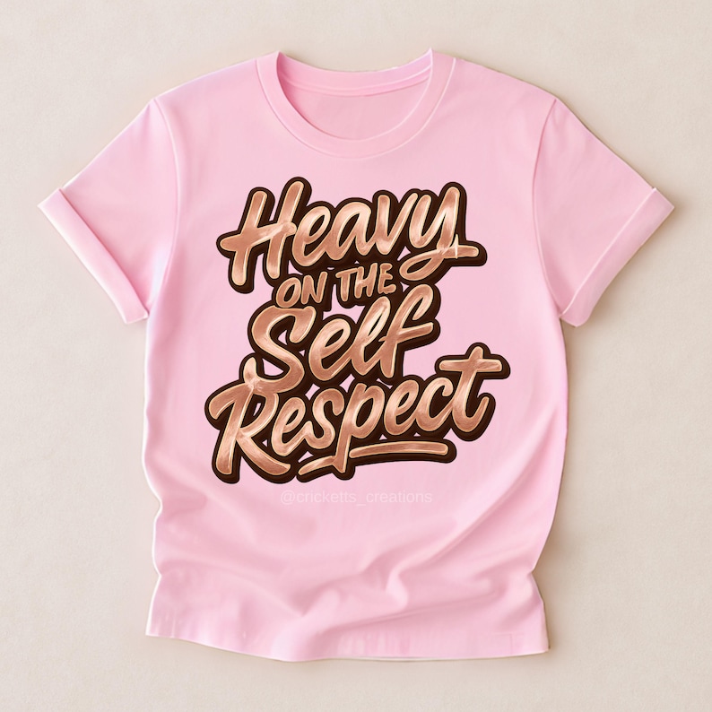 Puede incluir: Camiseta rosa claro con mangas enrolladas. La camiseta presenta la frase "Heavy on the Self Respect" en una fuente marr&oacute;n estilizada con contorno dorado. El texto est&aacute; centrado en la parte delantera.