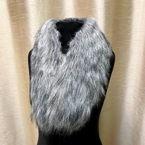 May include: A grey and white faux fur collar, presented on a black mannequin. The collar features a V-neck design and a soft, fluffy texture, providing a fashionable accessory.