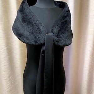 May include: A black faux fur shawl with a satin tie closure, displayed on a black mannequin. The shawl features a plush, textured fur collar and a smooth, flowing satin tie. The background is a neutral, light-coloured fabric.