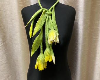 May include: A felted wool necklace in the shape of a flower arrangement. The necklace features long, green leaves and yellow and white flower buds. The necklace is displayed on a black mannequin.