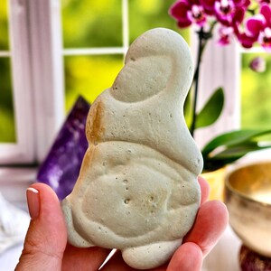 May include: A light green, seated Buddha statue. The statue is held in a hand, with a blurred background of a window, purple crystal, and a pink orchid. The Buddha has a smooth, rounded form with subtle details.