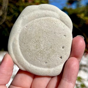 May include: A light gray, round, stone coaster with a textured surface and a raised edge. The coaster has a circular indentation in the center and is held in a hand.