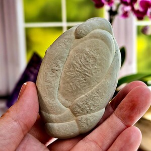 May include: A light gray, textured stone sculpture with an organic, rounded shape. The sculpture is held in a person's hand, with a blurred background of green foliage and purple flowers.