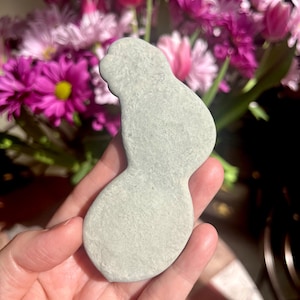 May include: A smooth, light green, irregularly shaped stone resembling a stylized human figure. The stone is being held in a hand.