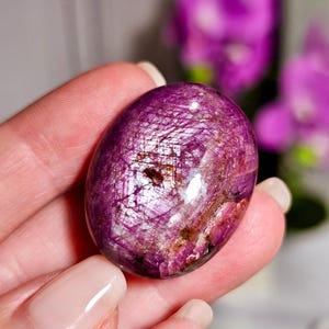 May include: A polished, oval gemstone in a deep magenta colour, featuring shimmering inclusions. The surface of the stone shows a network of fine lines and a glossy finish, indicating a high-quality mineral. The background is blurred, with hints of purple flowers.