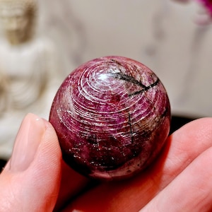 May include: A polished, spherical ruby gemstone featuring deep magenta and dark red hues with visible natural inclusions, dark mineral veins, and concentric surface patterns that reflect light across its rounded, smooth, and glossy exterior.