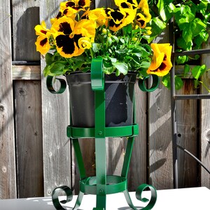 May include: A green metal plant stand with a black planter pot filled with yellow and brown pansies. The stand has a decorative scroll design and is sitting on a white surface.