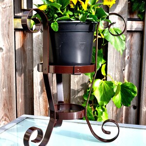 May include: A brown metal plant stand with a decorative scroll design. The stand has two tiers and is holding a black pot with yellow flowers.