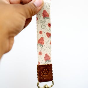 May include: A floral wrist strap with a cream background and red strawberry pattern. The strap has a brown leather accent with a flower design and a gold-colored clasp.