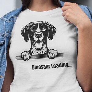 May include: White t-shirt featuring a black and white dog graphic with the text "Dinosaur Loading...". The dog illustration has a friendly expression. The shirt is worn with a blue denim jacket.