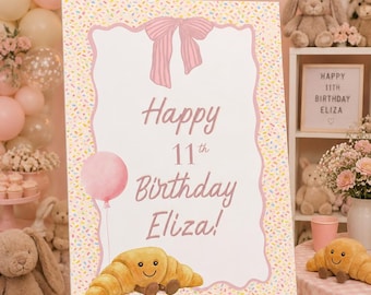 Puede incluir: Un cartel de cumpleaños rectangular sobre un caballete blanco con un borde de chispas de colores, un lazo rosa, un globo rosa y una ilustración de un cruasán sonriente. El texto dice Happy 11th Birthday Eliza! en una fuente cursiva rosa.