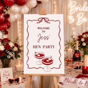 Puede incluir: Un letrero que dice "Welcome to Jessi Hen Party" se muestra en un caballete, con un borde rojo e ilustraciones de c&oacute;cteles. La escena incluye globos, flores, velas y una banda de "Bride to Be".