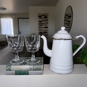 May include: Two clear glass stemmed drinking glasses with a decorative pattern sit on top of two vintage green and white books. A white enamel teapot with a black handle sits on a white surface.