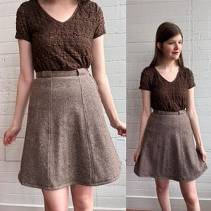May include: A brown textured V-neck top paired with a tweed skirt. The skirt is a light brown color with a subtle speckled pattern and a fitted waist. The top has short sleeves and a textured, crinkled appearance.