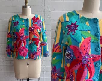 May include: A colorful, floral-embroidered cardigan with a round neckline and button closure. The sweater features a striped yellow and teal background with large, vibrant flower appliqués in shades of pink, red, orange, and purple. The garment is displayed on a mannequin.