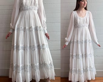 May include: A white maxi dress with long, sheer sleeves and a tiered skirt. The dress features delicate blue floral embroidery along the bodice and skirt tiers. The dress is paired with cream-colored pointed-toe heels.