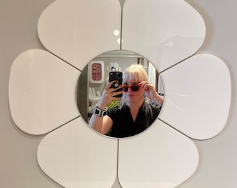 Puede incluir: Un espejo blanco con forma de flor de siete pétalos. Una persona se refleja en el pétalo central, llevando gafas de sol y sosteniendo un teléfono.