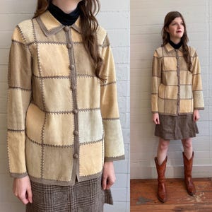 May include: A long-sleeve jacket constructed from square patches of tan and beige suede leather. The garment features a collared neckline, a front button closure with matching suede buttons, and decorative whipstitch detailing along the seams.